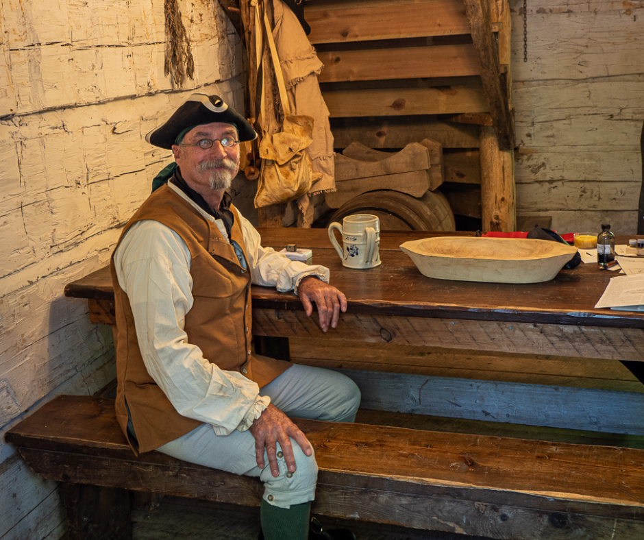 Man in 18th century clothing sitting at a table in rustic historic cabin