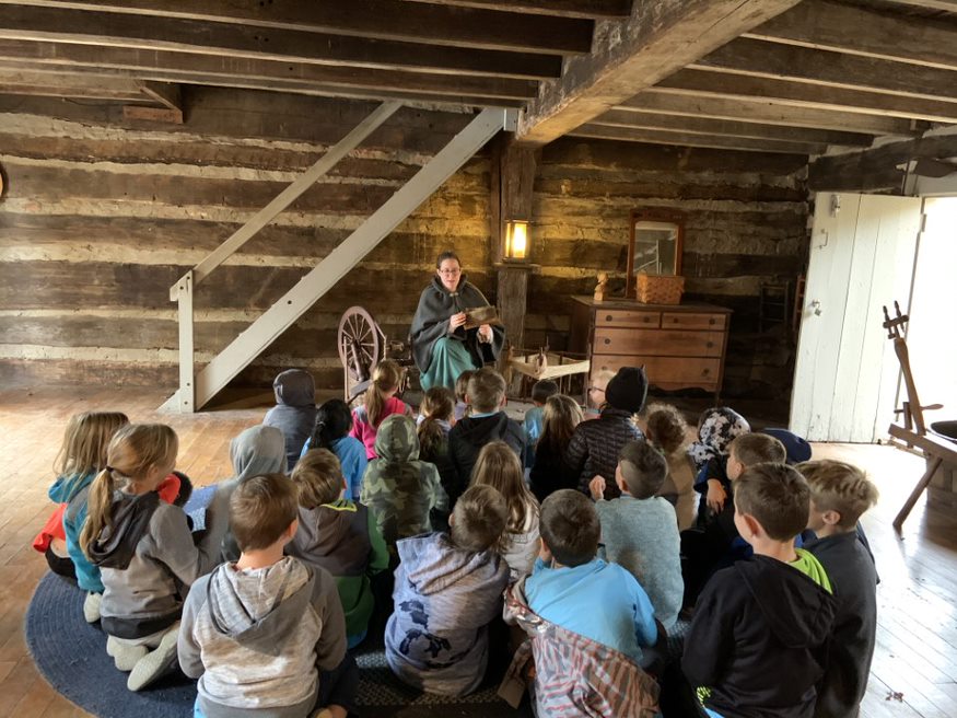 Students sit on floor in cabin while educator dressed in pioneer clothing sits at spinning wheel